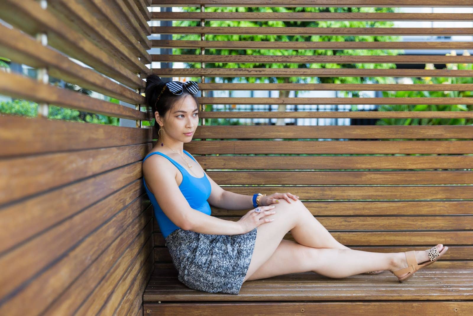 A woman with dark hair in a bun, wearing a blue bodysuit and a black and white textured skirt. She is sitting on a wooden bench in a cabana, with her legs outstretched and ankles crossed. She is wearing nude coloured sandals with a spotted strap over the toes