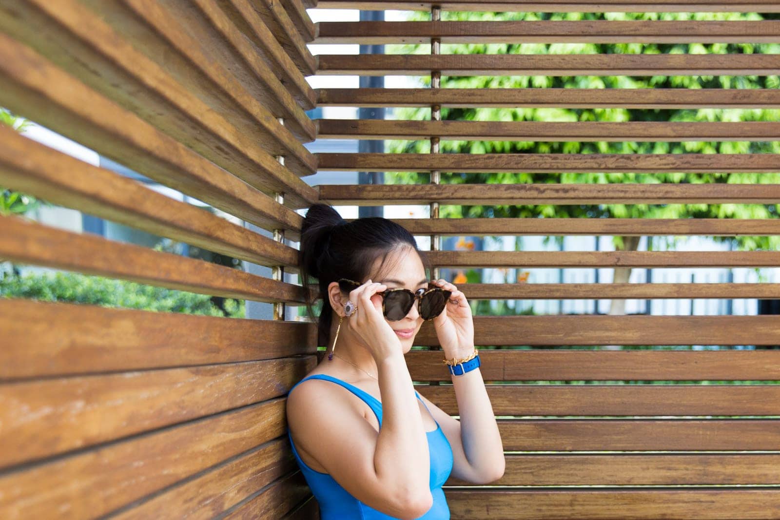 A woman with dark hair in a bun, wearing a blue top. She is holding her round sunglasses to her face, almost covering her eyes. Her eyes are wide open