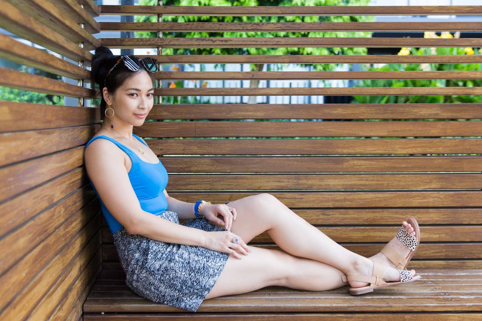 A woman sitting in a wooden cabana, she is wearing a blue sleeveless bodysuit and a black and white textured skirt. Her gaze is away from the camera.