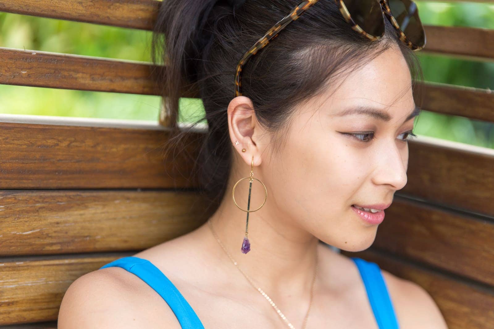 A closeup of a woman’s face, her face angled slightly away from the camera. She has gold hoop earrings in her ear with a raw amethyst crystal drop