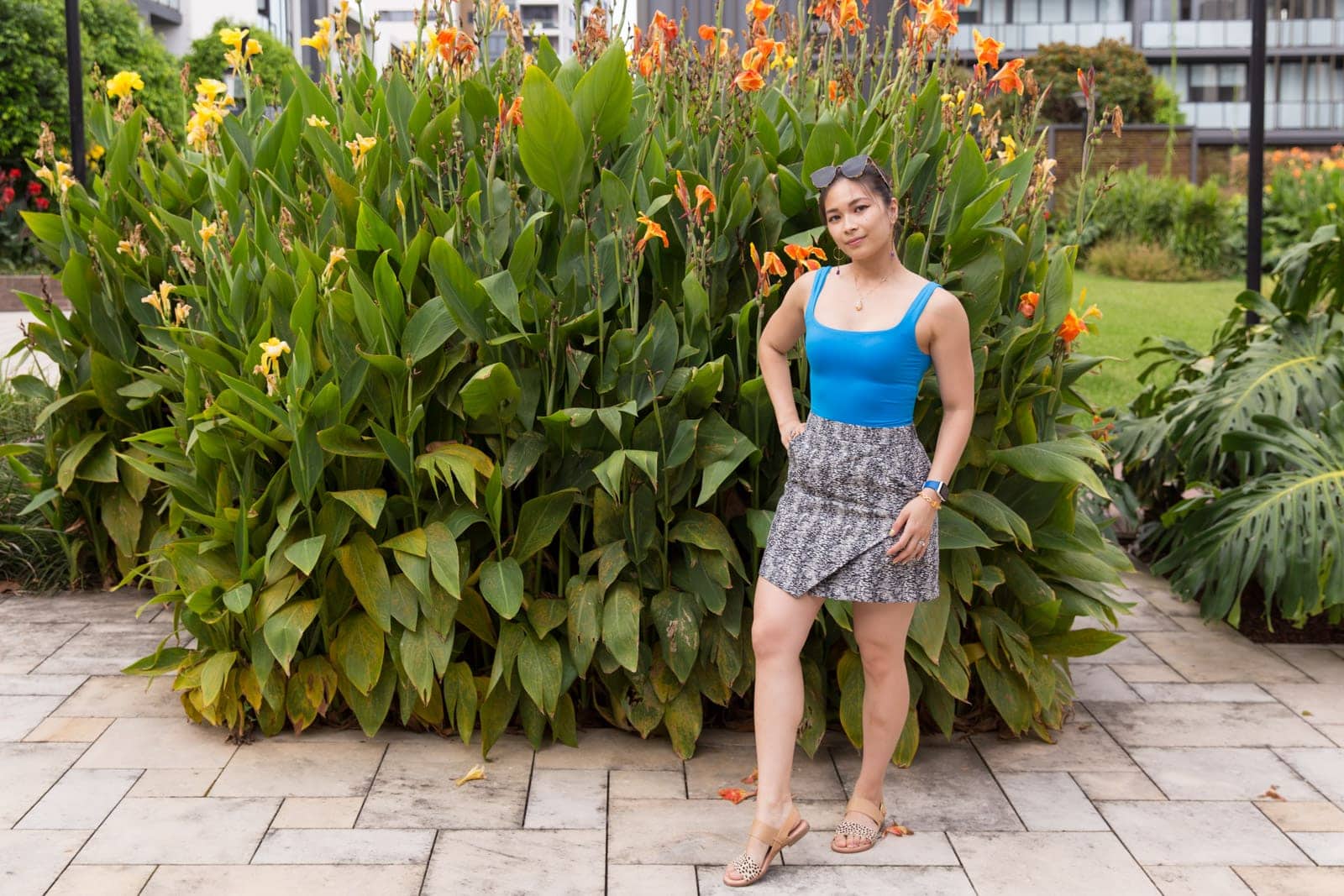 A woman in a blue bodysuit and black and white textured skirt, in front of a large planting of tall plants. She has sunglasses on top of her head