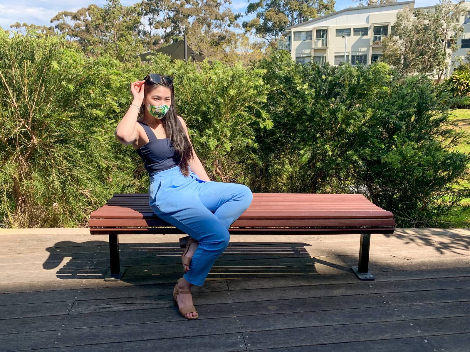 The same woman in other photos on this page, wearing the same clothes. She is sitting on a wooden bench, leaning back a little, with her ankles crossed at the ground beneath the bench. She is holding onto her sunglasses on top of her head