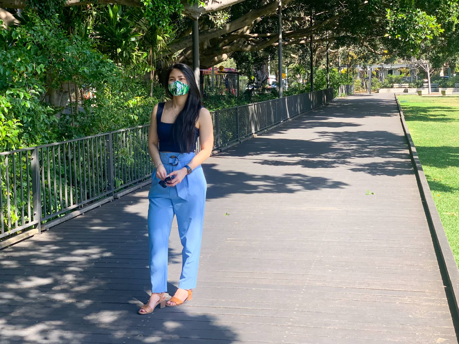 A woman with long dark hair, wearing a navy blue square neck top and a sky blue pair of pants with a buckle detail at the waist. She is wearing a face mask with a green leaf pattern. She is holding sunglasses in her hands and is standing on a wooden path.