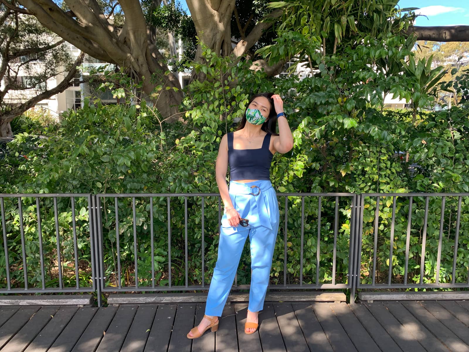 The same woman in other photos on this page, wearing the same clothes. She is standing in front of a metal fence and one of her hands is brushing her hair back on top of her head. In the other hand she is holding a a pair of sunglasses