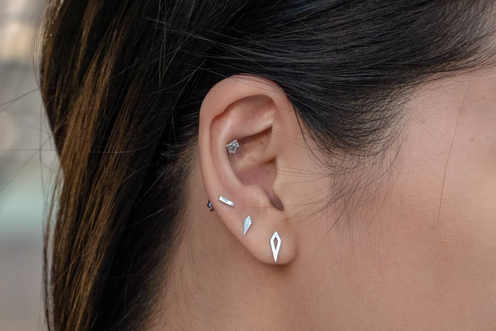 Close up of a woman’s ear with three small silver lobe earrings and one small cartilage piercing.