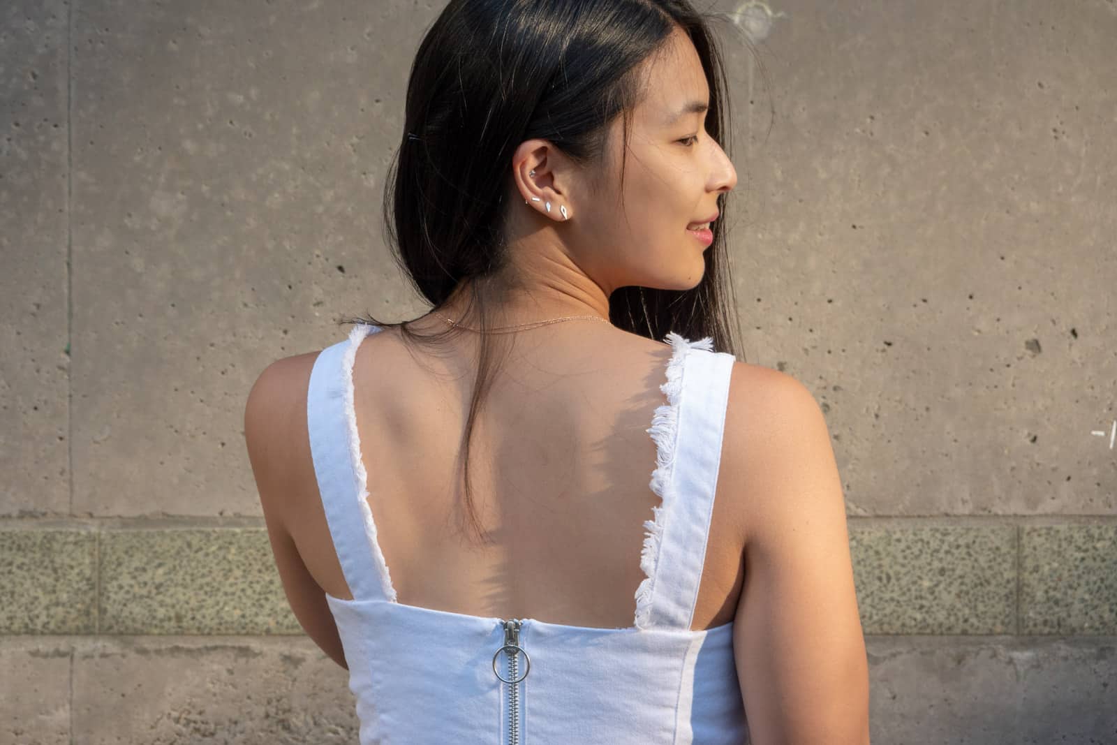 From behind, a woman facing the right, with the O-ring pull zipper of her strapless white top visible. Some of the woman’s dark hair falls across her back