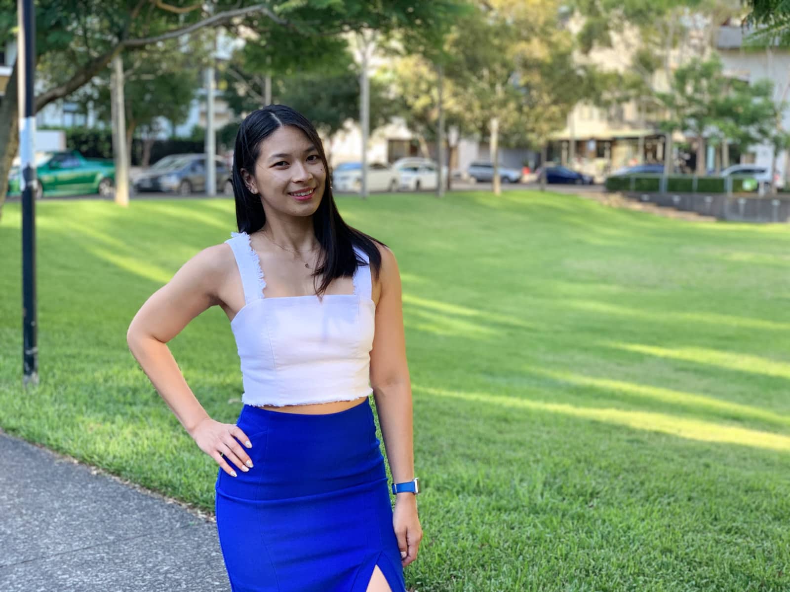 A woman with short dark hair in a white strapless top and a cobalt blue skirt, with a hand on her hip. There is a park with green grass behind her