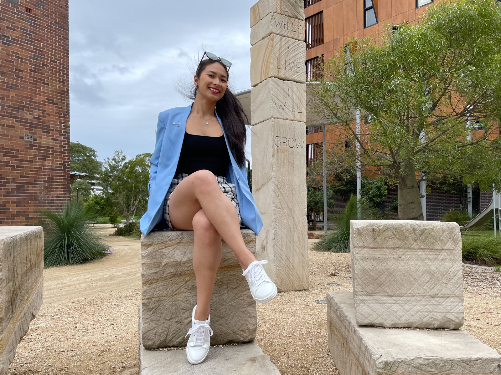 An Asian woman with long dark hair tied in a ponytail. She is wearing checkered shorts and a blue blazer. She is sitting on a giant stone step, next to a pillar carved with “While I live, I will grow”.