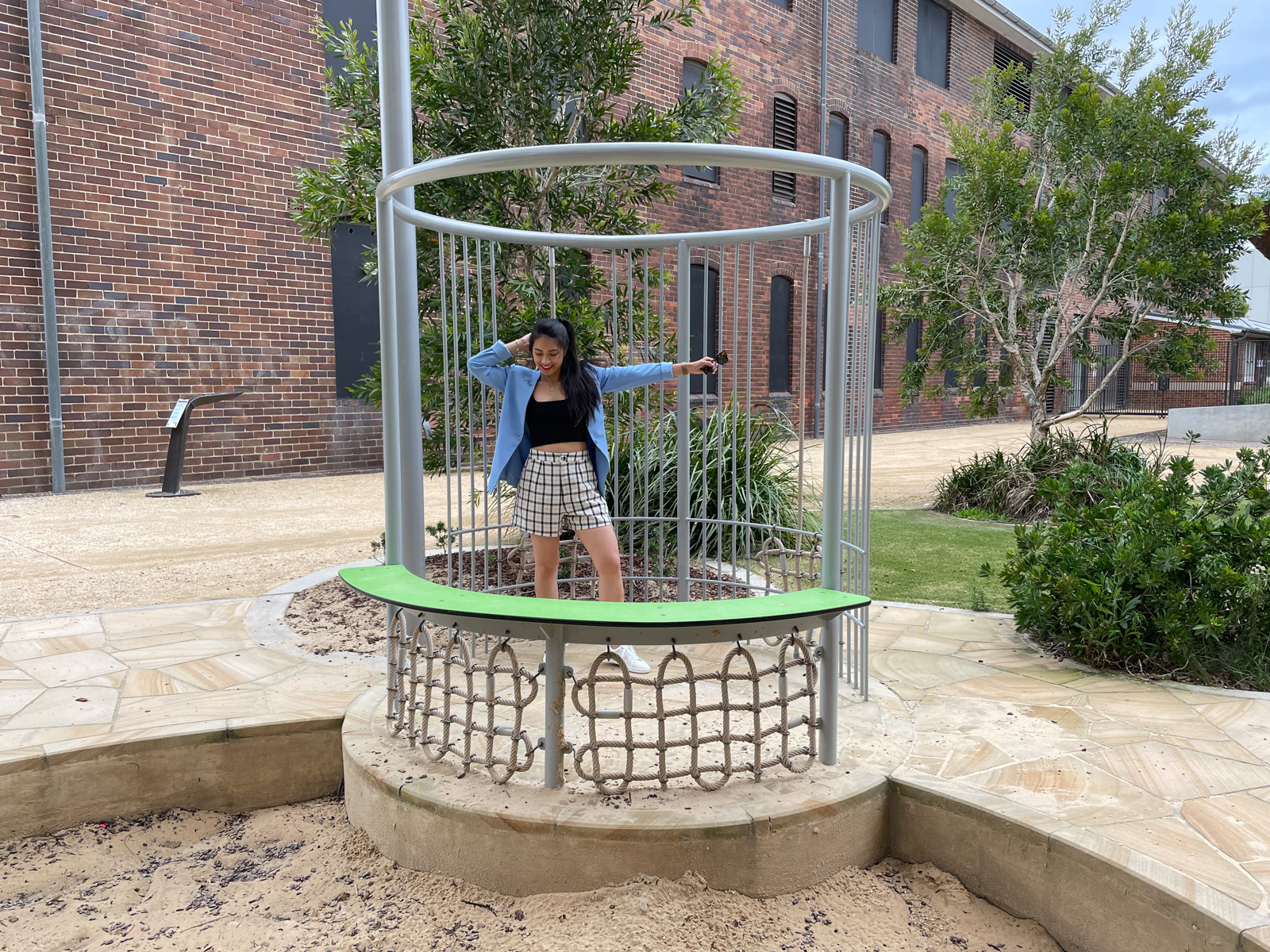 The same woman from previous photos on this page, wearing the same outfit. She has a hand behind her head and the other arm outstretched, looking down slightly. She is standing in a small metal structure right next to a sandpit