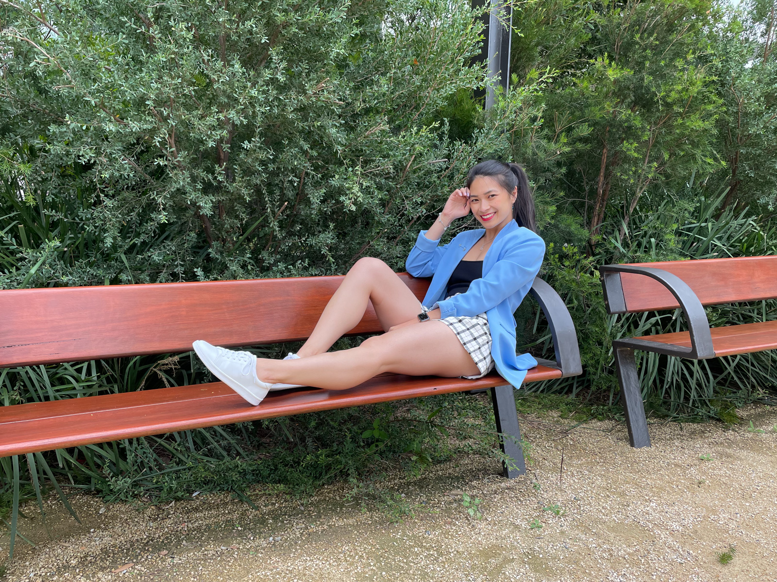 The same woman from previous photos on this page, wearing the same outfit. She is sitting across a bench with one of her legs outstretched and the other with the knee bent. She has one hand in her lap and the other at the side of her face with her elbow bent.