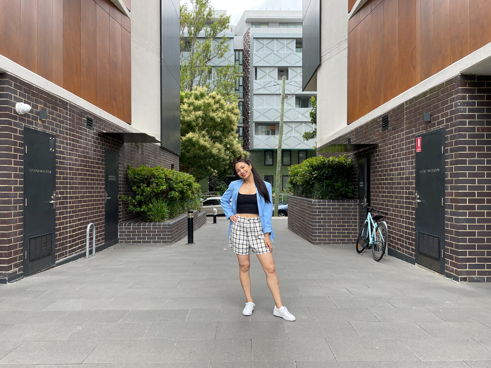 The same woman from previous photos on this page, wearing the same outfit. She is standing in a walkway with brick walls with storeroom doors on either side. There is a street and apartment building in the background behind her. She has one hand on her hip.