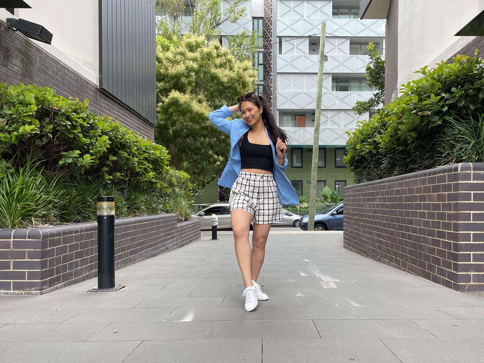 A woman posing in a walkway between two brick walls with plants in them. Some apartment buildings are in the background. The woman is wearing a blue blazer over a black crop top and black-and-white plaid shorts. She has white sneakers on. Her long, dark hair is in a ponytail which she is holding the end of with one hand. Her other arm is bent at the elbow with her hand behind her head. One of her knees is bent.