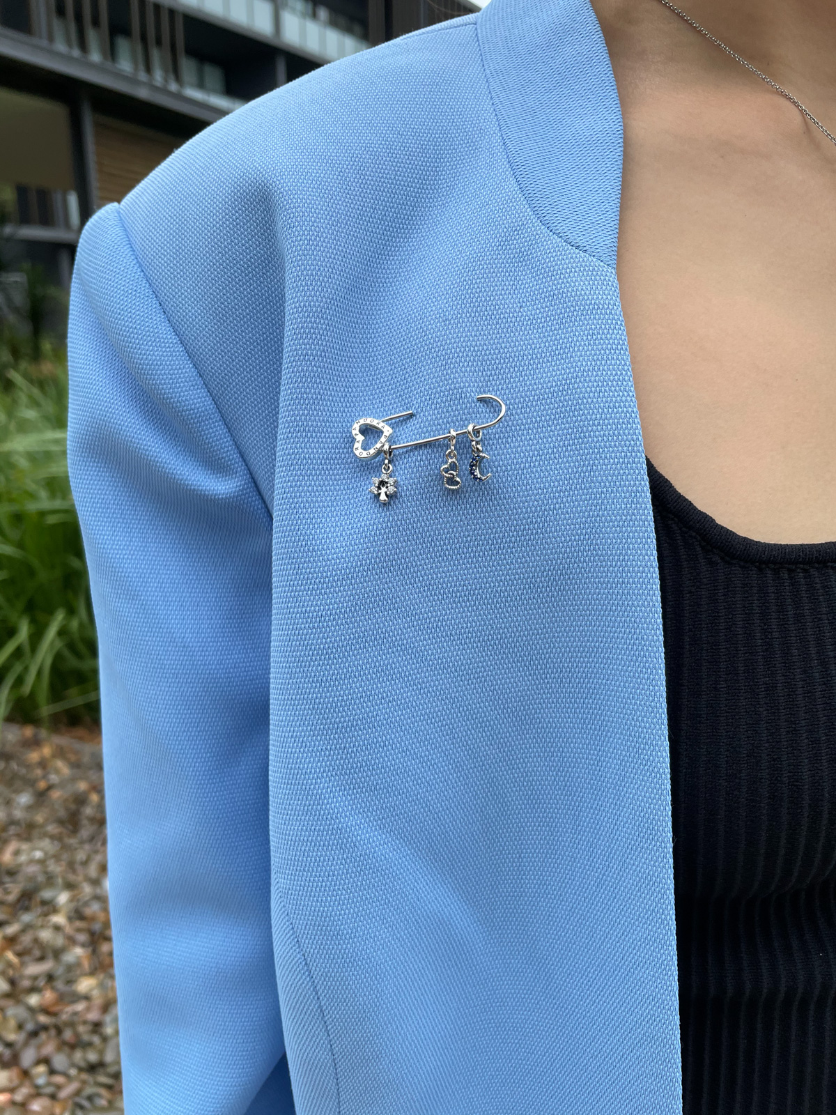 A close-up of the lapel of a woman’s blue blazer. It has an adorned safety pin with three charms pinned on it: a tree; two hearts; and a moon. The tree and moon are adorned with rhinestones