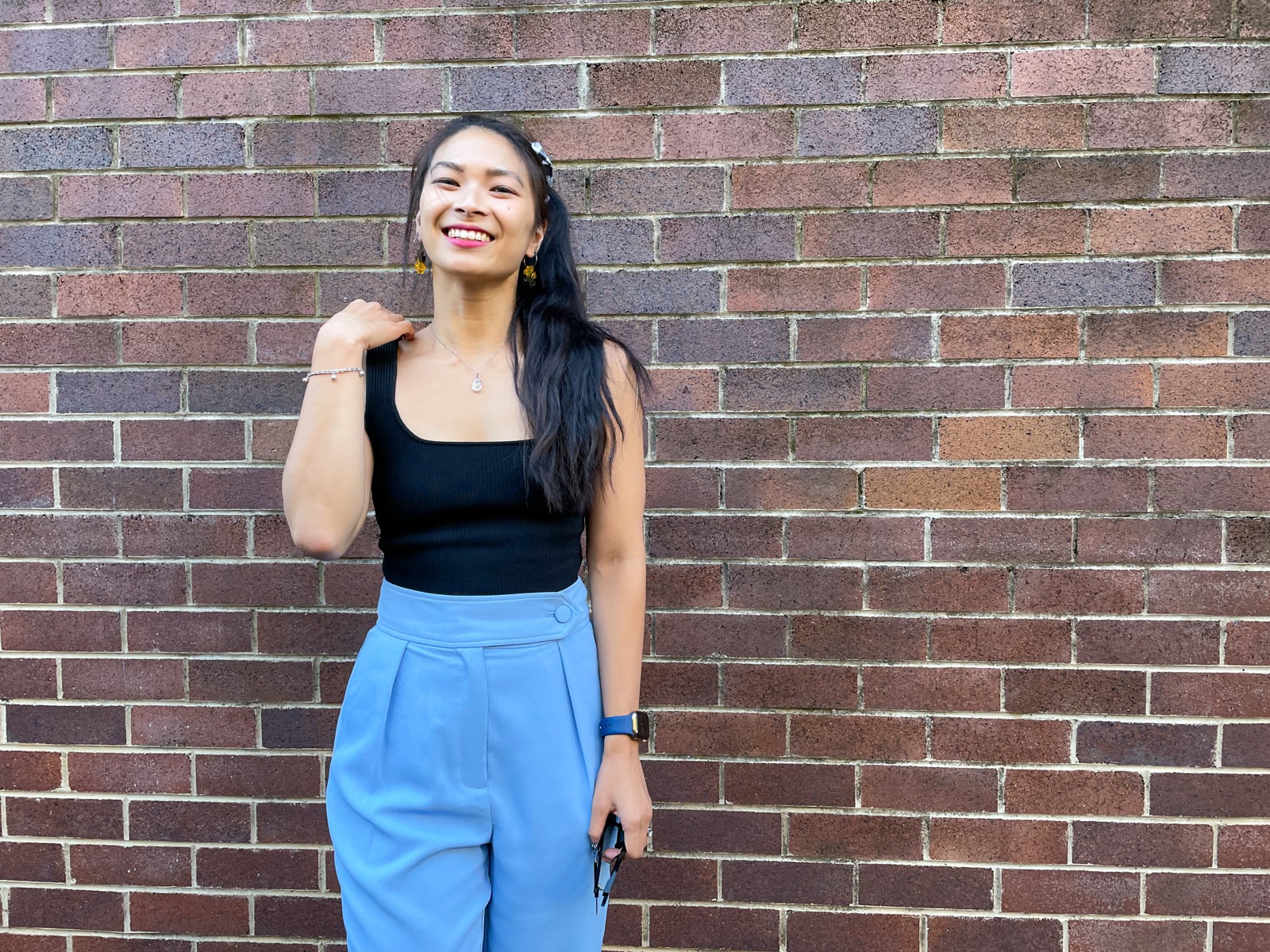 An Asian woman wearing a black square neck sleeveless top and blue trouser pants. She has her long dark hair in a ponytail and is holding a pair of sunglasses. Her other arm is bent and touching her shoulder. She is standing in front of a brick wall and smiling.