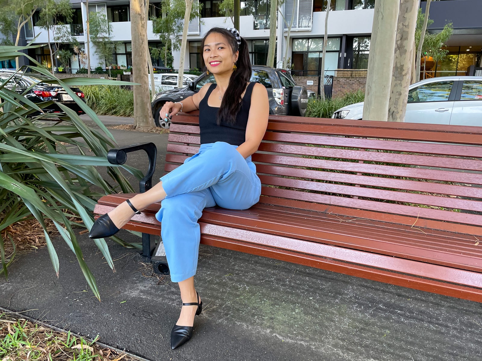 An Asian woman wearing a black square neck sleeveless top, sky blue trouser pants, and pointed black sandals. She is sitting on a wooden park bench and has one leg crossed over the other, and an arm leaning on the back of the bench.