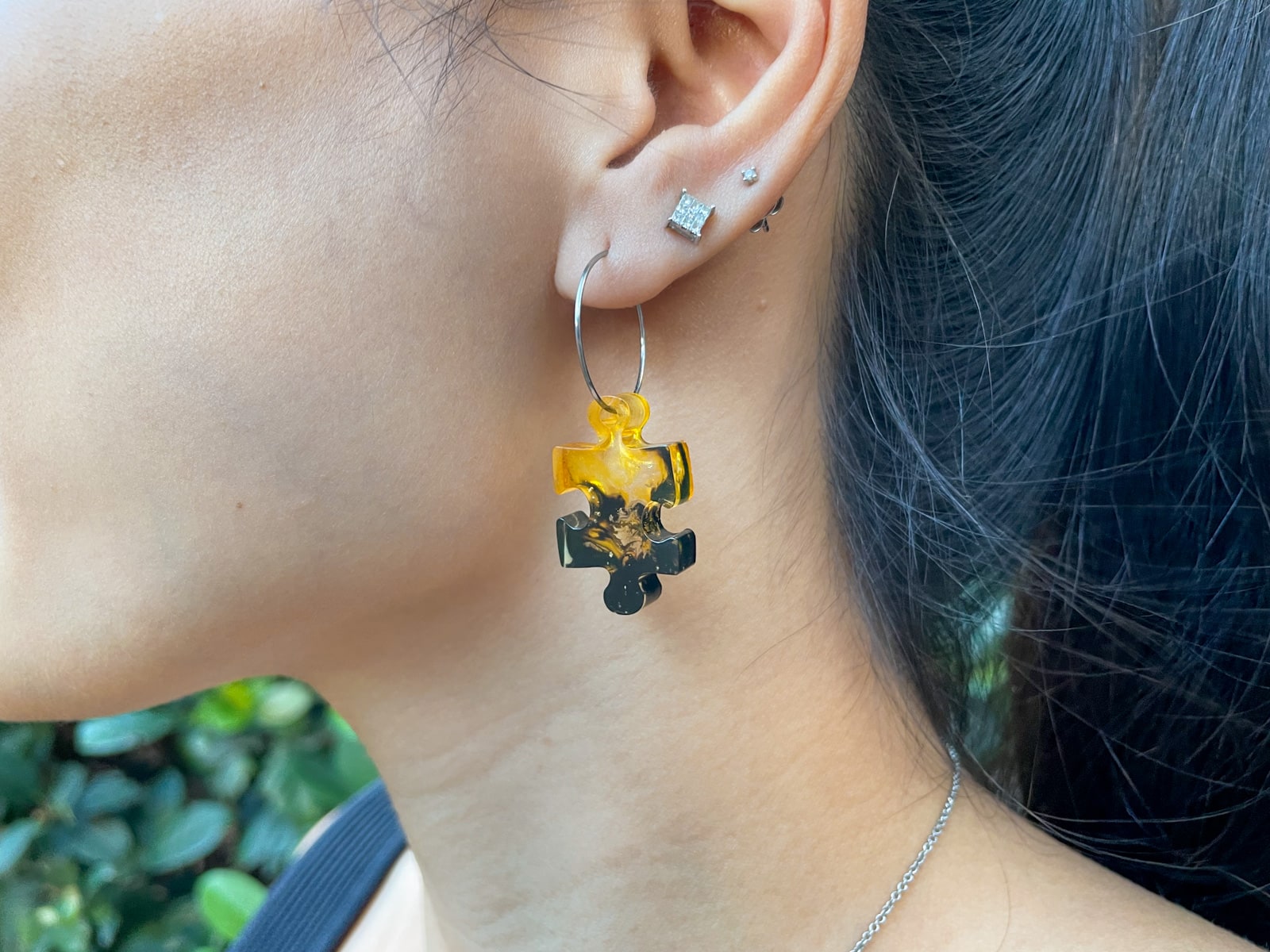 A close-up of a womanâ€™s ear with two silver earrings in the upper lobe, and a silver hoop earring with a yellow and black puzzle-shaped resin drop in the lower lobe.