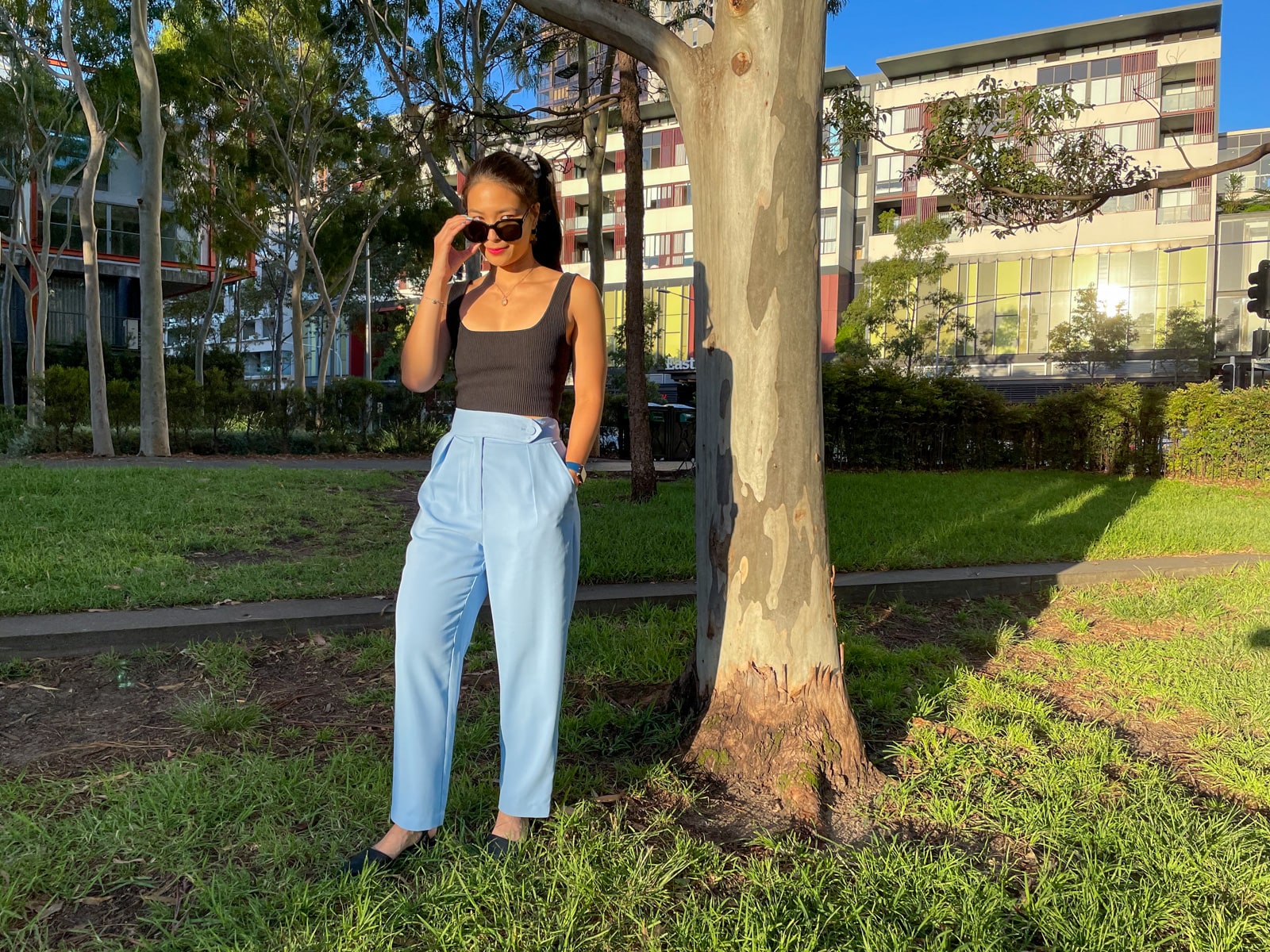 An Asian woman wearing a black square neck sleeveless top and sky blue trouser pants. she is standing beside a tree trunk, and has her hand held up to her black sunglasses on her face. The sun indicates it’s late afternoon.