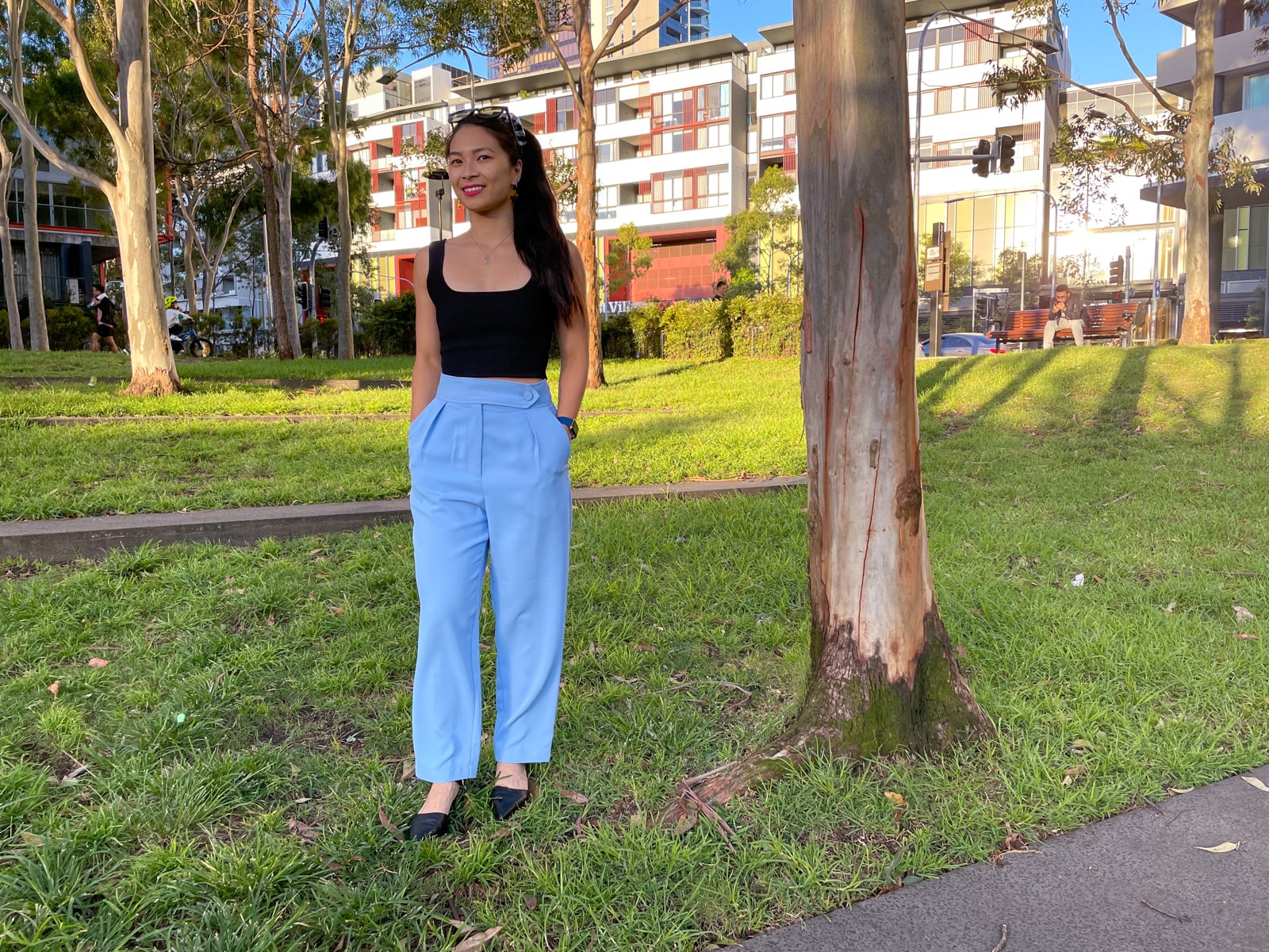 An Asian woman wearing a black square neck sleeveless top, sky blue trouser pants, and pointed black sandals. She is standing next to a tree in a park, with her hands in her pockets.