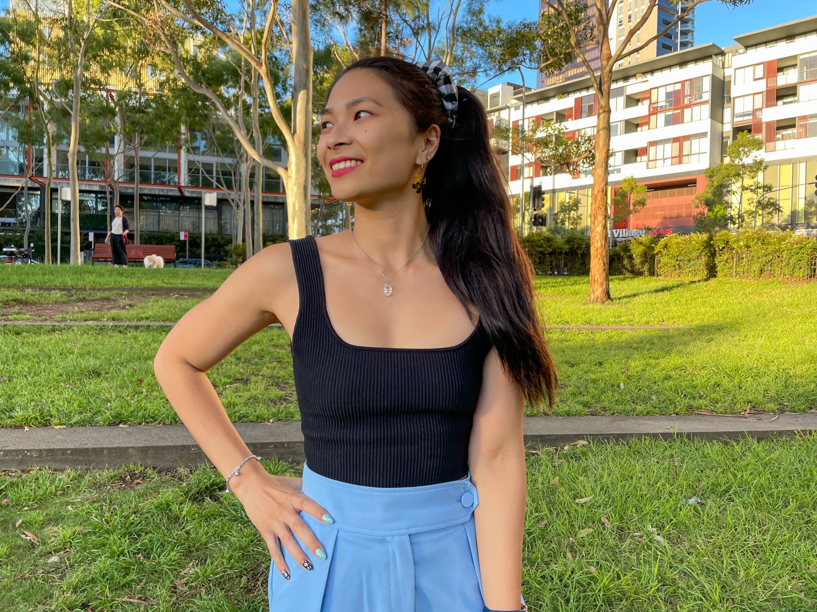 An Asian woman wearing a black square neck sleeveless top and sky blue trouser pants. She has a hand on her right hip, and is looking to her right. Her long dark hair is in a ponytail with a black and white gingham printed scrunchie.