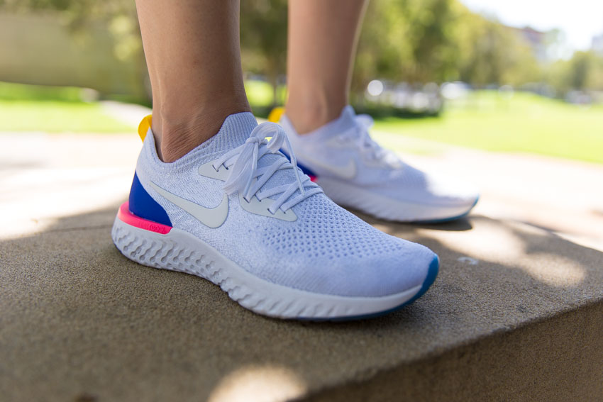 A white running shoe with white laces on a person’s right foot. It is accented by a small panel of bright blue and hot pink at the base of the heel, and a bright yellow tab behind the top back of the shoe. The front toe of the shoe shows a bit of blue. The person’s left foot wearing the matching shoe is in the background.