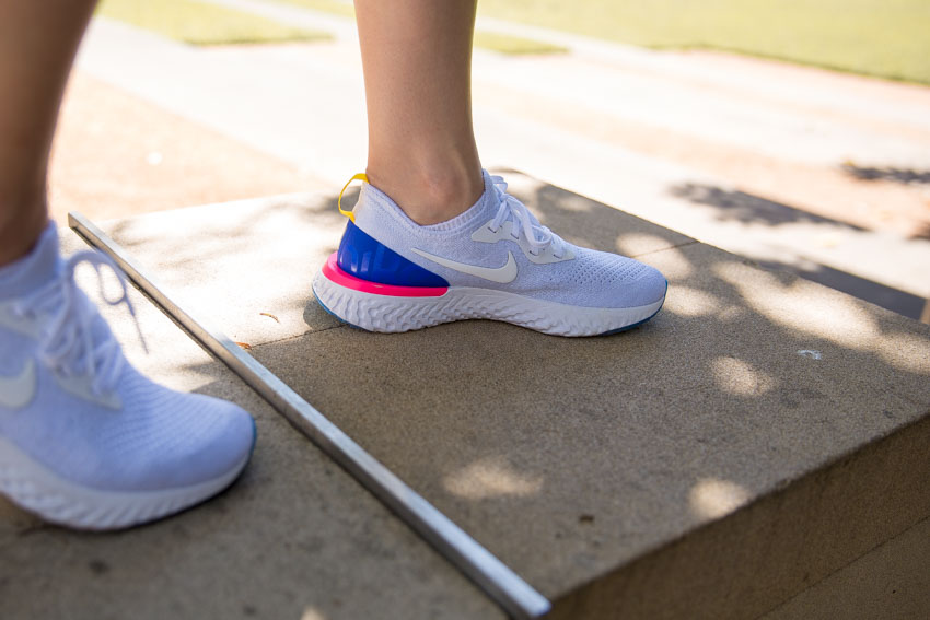 A white running shoe with white laces on a person’s left foot. This supplements the previous picture. The shoe is accented by a small panel of bright blue and hot pink at the base of the heel, and a bright yellow tab behind the top back of the shoe. On this shoe, the brand Nike’s swoosh logo is visible on the inner side of the shoe. It has enough contrast to the rest of the shoe to stand out subtly.