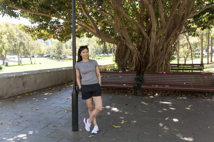 Me, smiling, leaning against a black lamppost on my right-hand-side. The top of the lamppost is not visible. My right ankle is crossed in front of my left and I have my hands in the pockets of my black shorts. I am wearing a grey t-shirt and have sunglasses on top of my head. There is a brown wooden bench in the background, as well as a large tree with large branches.