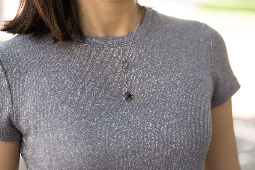 A close shot of my t-shirt at the neck. A thin silver chain sits around my neck and joins to a silver ring, which has a green and purple ruby drop pendant hanging from a chain downwards from the ring.