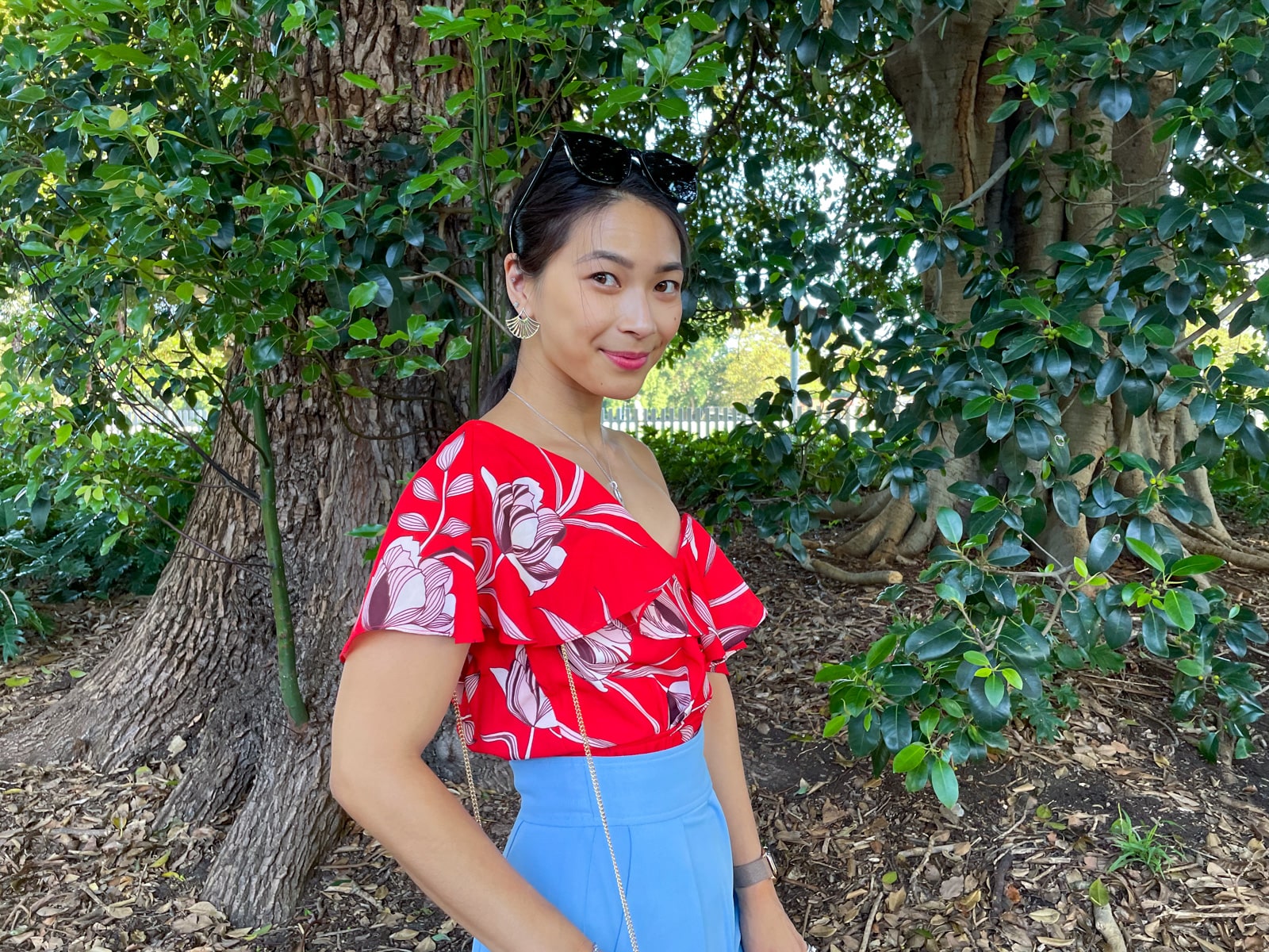 An Asian woman wearing a red top with a floral print, and blue pants, with her hair tied back in a low ponytail. She has black sunglasses on top of her head and is smiling with her chin pointed downwards as she looks up. She is standing in front of a tree.