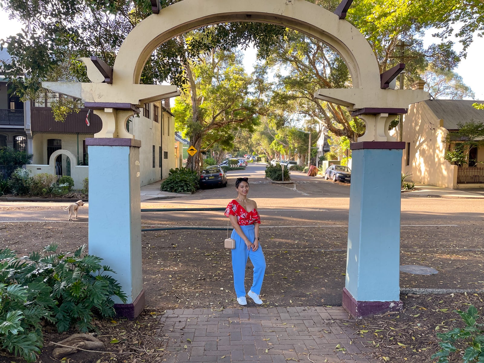 An Asian woman wearing a red top with a floral print, and blue pants, with her hair tied back in a low ponytail. She has white shoes on. One of her knees is bent and her hands are clasped together. She’s standing under a concrete arch that marks the entrance to a park. Behind her is a suburban street.