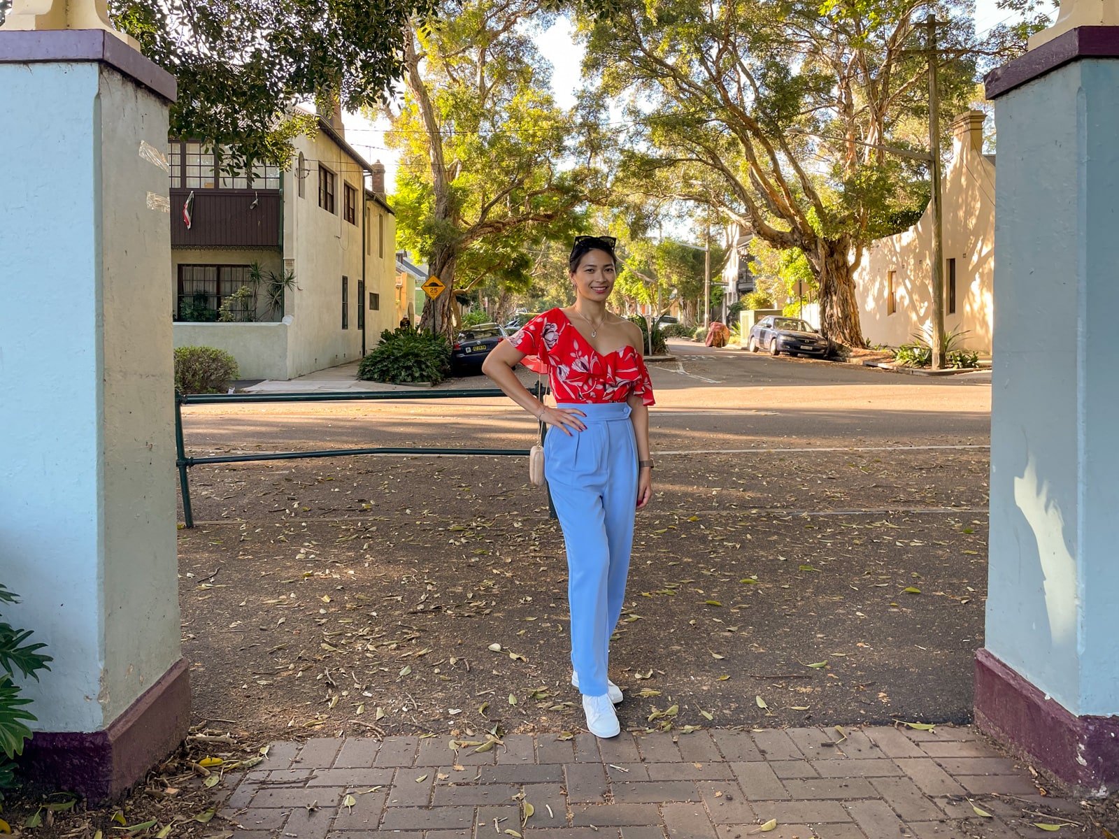 An Asian woman wearing a red top with a floral print, and blue pants, with her hair tied back in a low ponytail. She has a hand on her hip, showing a gold coloured rounded rectangular clutch bag hanging from a chain on her shoulder. She is standing under a concrete arch at a park.