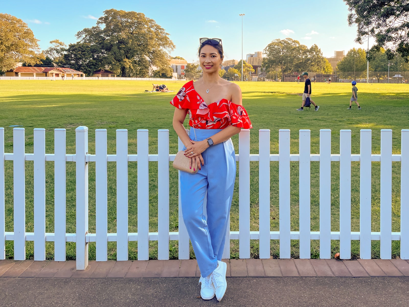 An Asian woman wearing a red top with a floral print, and blue pants, with her hair tied back in a low ponytail. She has black sunglasses on top of her head and is wearing white shoes. She holds onto a gold clutch bag hanging from her shoulder. She stands in front of a white fence along a sports oval.