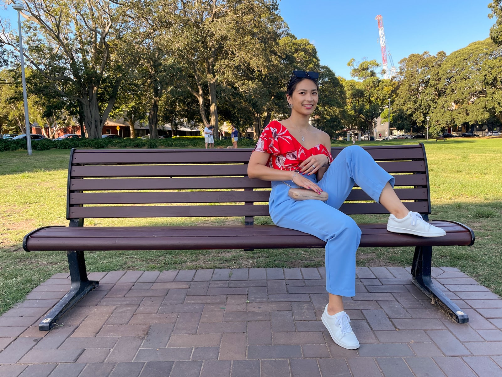 An Asian woman wearing a red top with a floral print, and blue pants, with her hair tied back in a low ponytail. She has white sneakers on and is sitting on a bench, with a leg bent and her foot up on the bench.