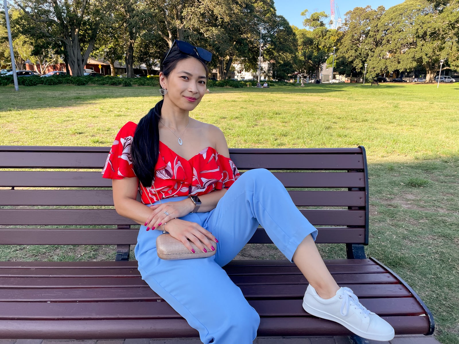 An Asian woman wearing a red top with a floral print, and blue pants, with her hair tied back in a low ponytail. She is leaning back on a bench with a gold clutch handbag in her lap. She has a foot up on the bench with her knee bent.