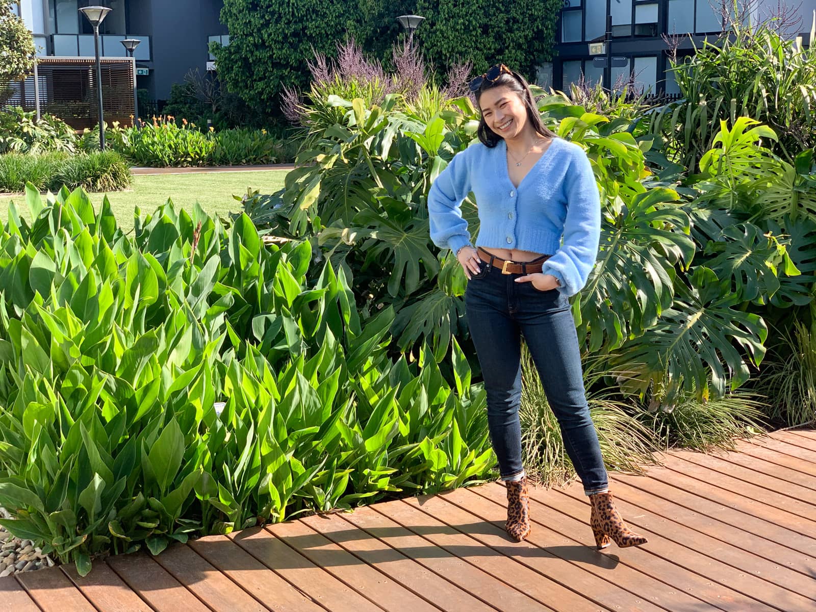 The same woman in the previous photo on this page, in the same outfit. She is standing on a wooden boardwalk with a hand on her hip and another in her jeans pocket. Behind her are some large green plants.