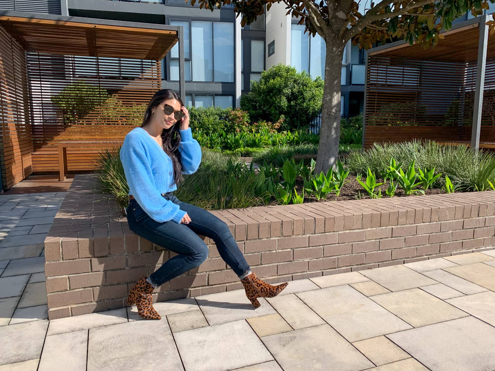 The same woman in the other photos on this page, in the same outfit. She is sitting on a short brick wall and has both her knees slightly bent. She has brown ankle boots on with a black leopard print. She has round tortoiseshell sunglasses over her eyes. In the background is a wooden cabana with a bench.