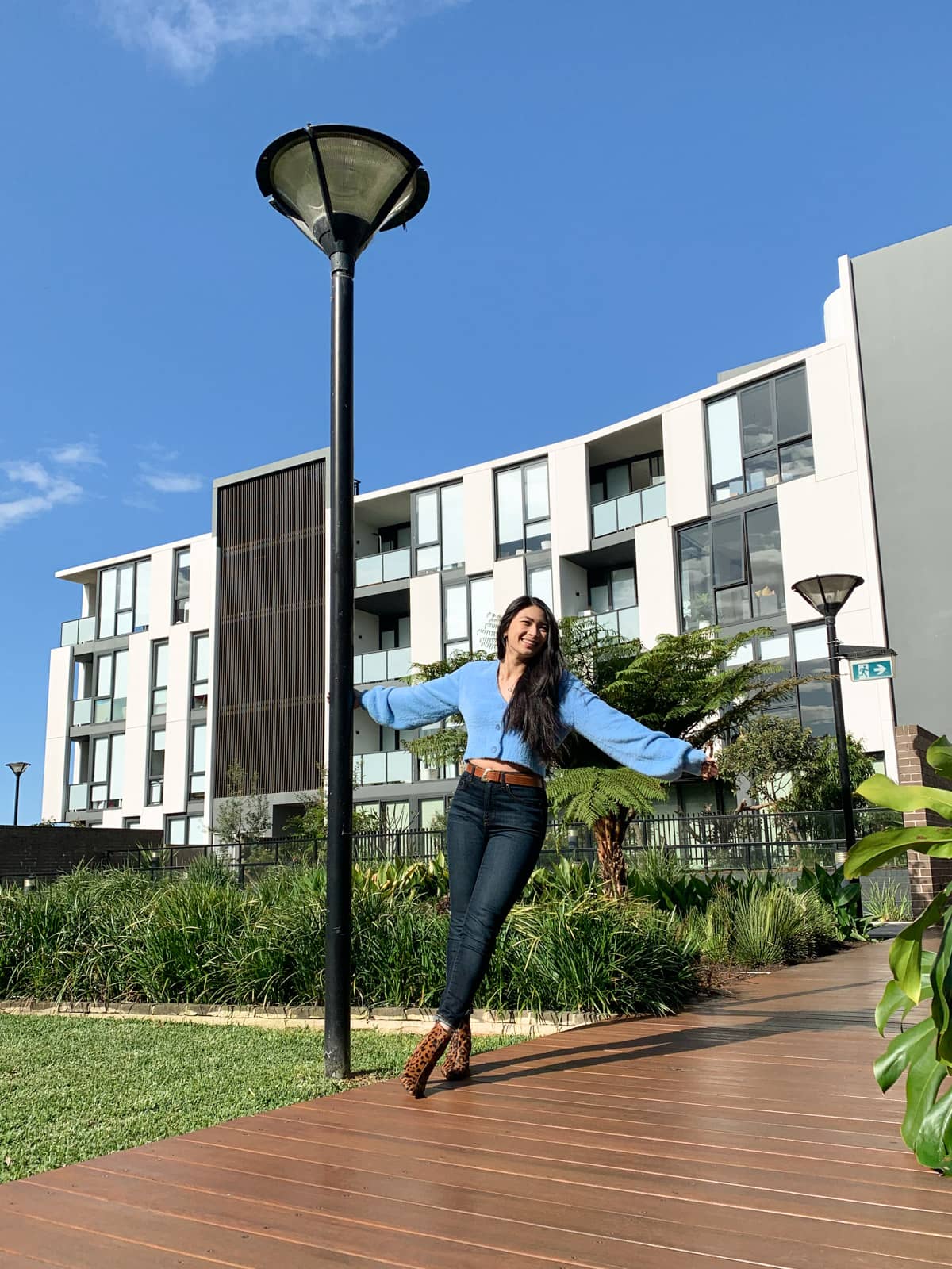 The same woman in the other photos on this page, in the same outfit. She is holding onto a lamppost and stretching out to the opposite side, with her ankles crossed. She is standing on a wooden boardwalk. In the background is an apartment building and the sky is blue like her cardigan.