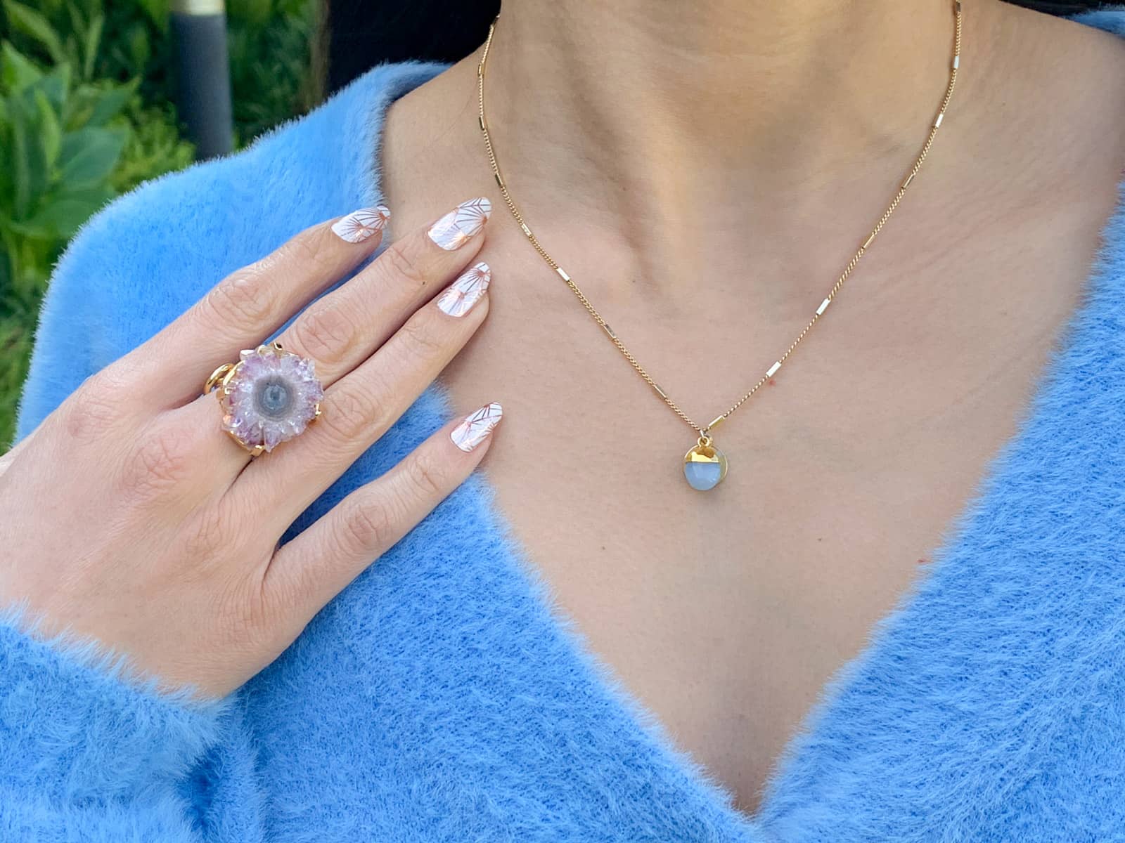 A close-up of a woman’s neckline; she is wearing a gold necklace with a blue gemstone that matches the blue cardigan she is wearing. Her hand is resting on her collarbone, and her fingernails have white nail art with rose gold sunburst patterns. She is wearing a raw amethyst ring on her middle finger.
