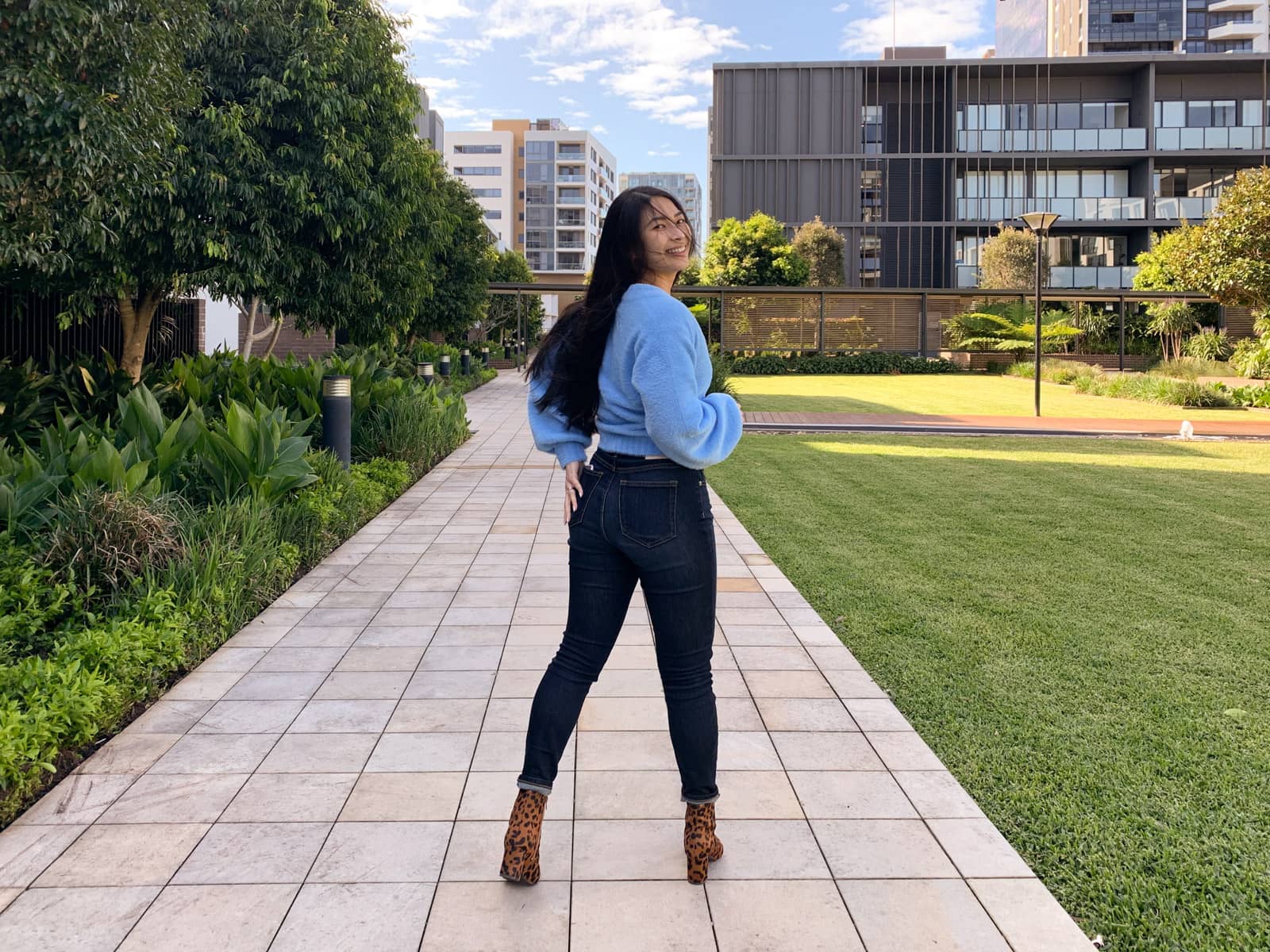 The same woman in the other photos on this page, in the same outfit. Her back is to the camera and one of her hands is reaching for her back jeans pocket, but she is turning to look over her right shoulder. In the background is an apartment building and a trimmed green lawn.