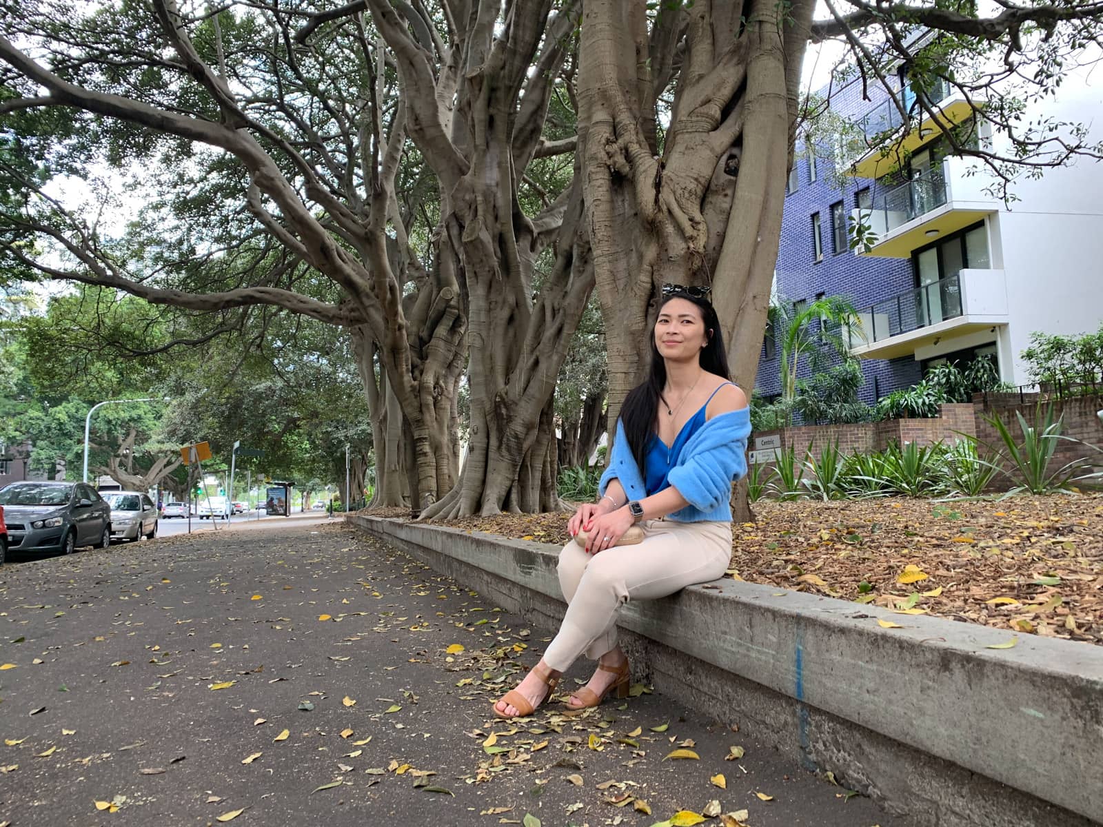 A woman with long dark hair sitting on a short wall at the end of a garden. She is wearing light pants, a blue top and a sky blue fluffy cardigan. In the background are apartment buildings and trees with large trunks.