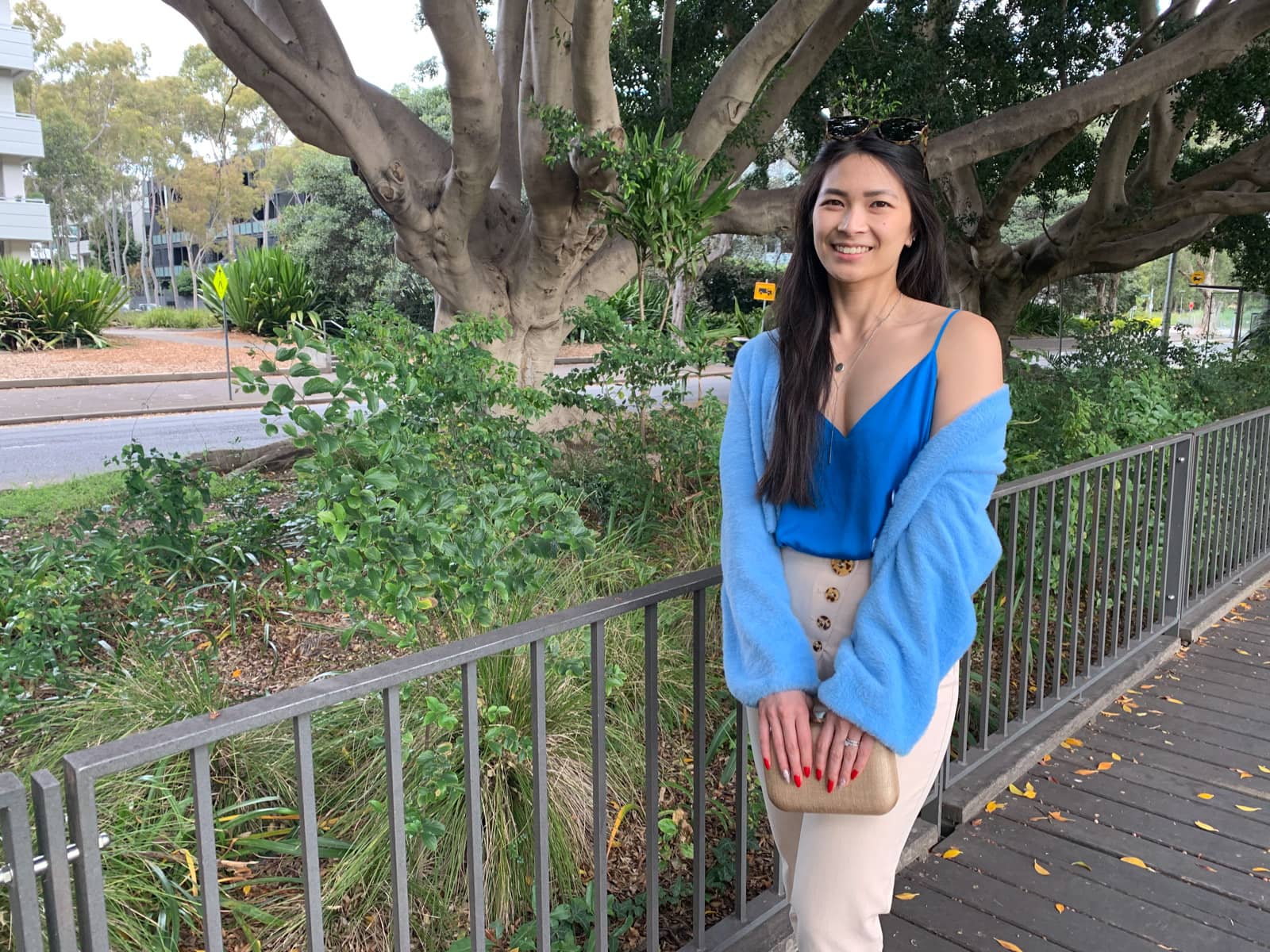 The same woman in the previous photo on this page, in the same outfit. She is standing on a wooden boardwalk, holding a rounded rectangular tan-coloured clutch with both hands. Her cardigan is lightly draped over her shoulders with one shoulder exposed. She is smiling widely.