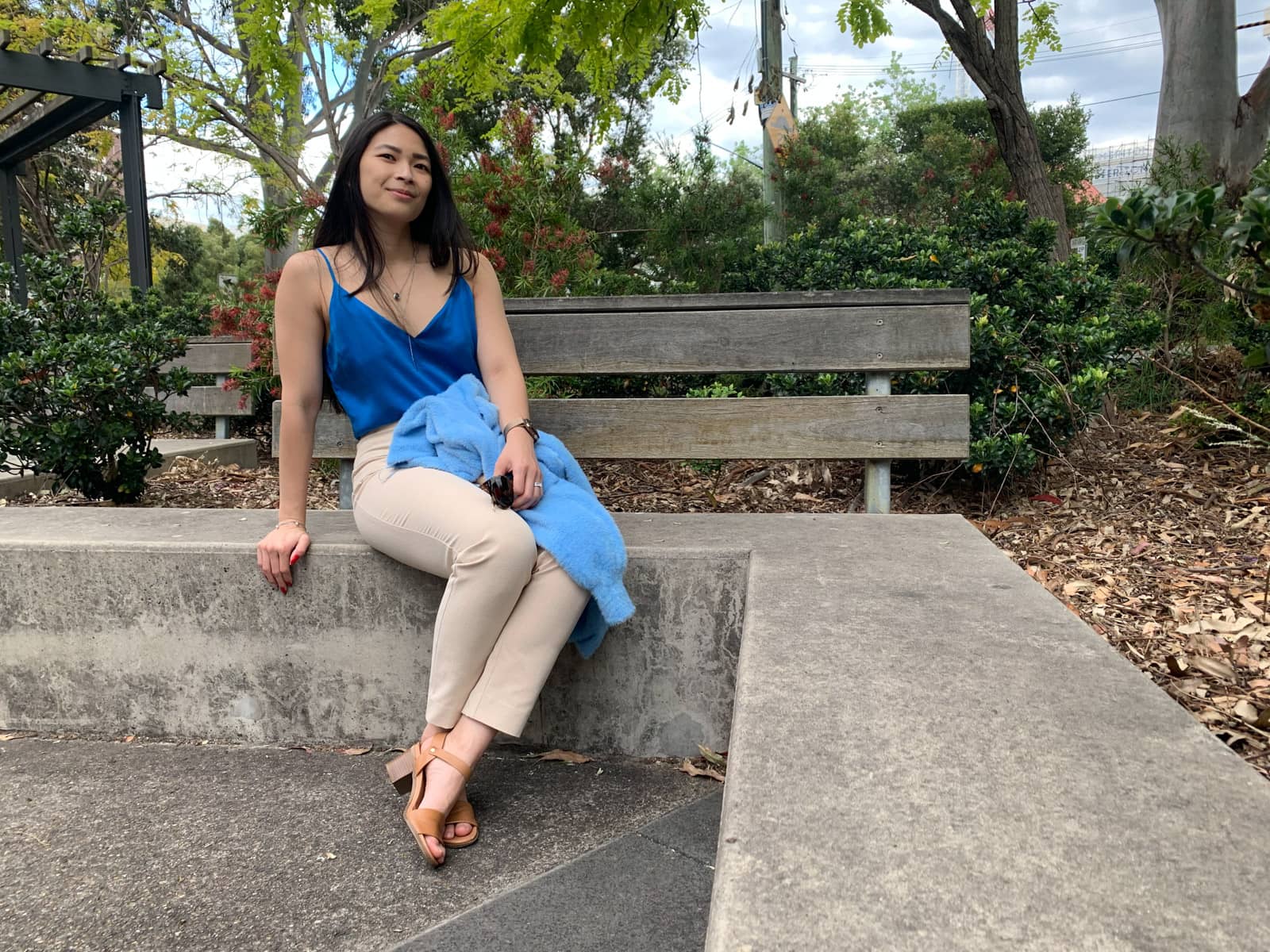 The same woman in the previous photos on this page, in the same outfit. She is sitting on a short concrete wall with both knees bent and her ankles crossed. She is wearing tan coloured sandals. She is leaning with one hand at her side for balance, and the other in her lap with a cardigan on her lap.