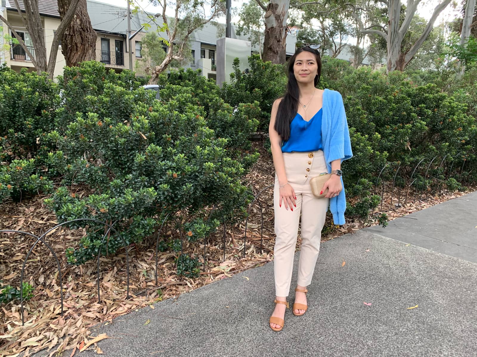 The same woman in the previous photos on this page, in the same outfit. She is standing in front of shrubbery and has one hand clutching her handbag and the other resting on her upper thigh