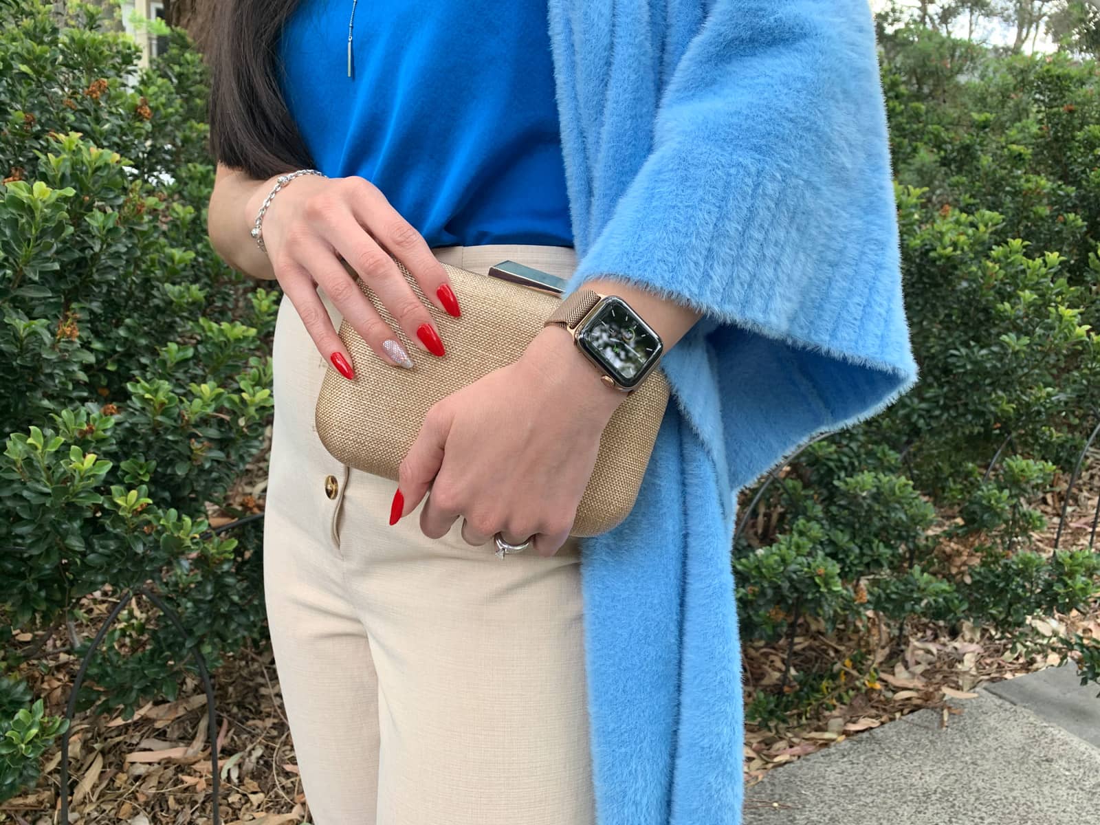 A close-up of a woman’s red manicured long nails clutching a small rounded rectangular clutch handbag. Her sky blue cardigan can be seen and she is wearing a bright blue top and light pants