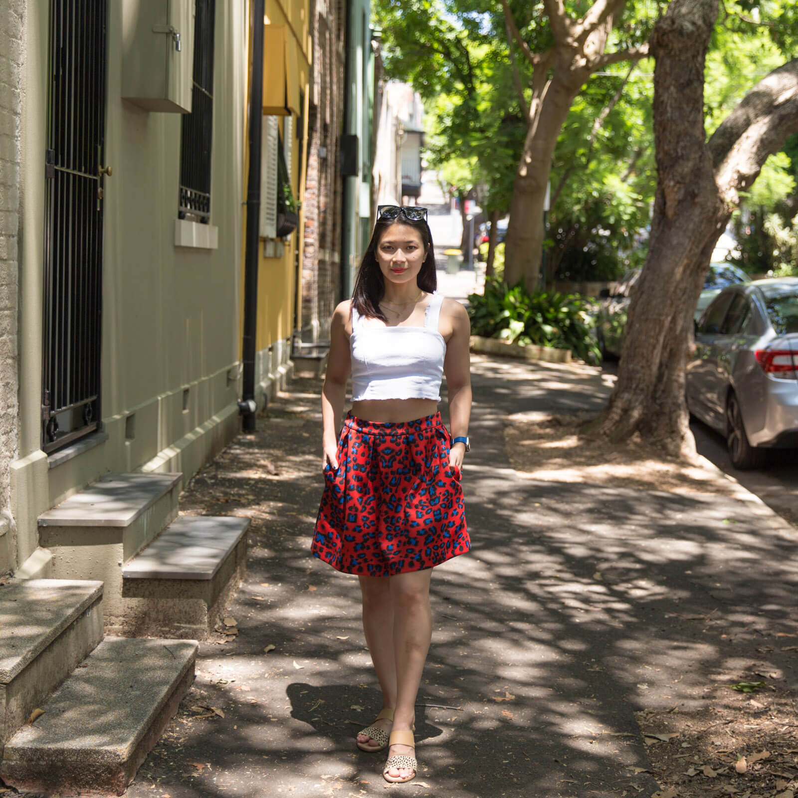 A woman standing on a footpath next to steps that lead up to the front door of houses. The area is shaded by trees. The woman has dark hair, and is wearing a white top with a red skirt with blue leopard sports. She has her hands slightly in the pockets of her skirt.