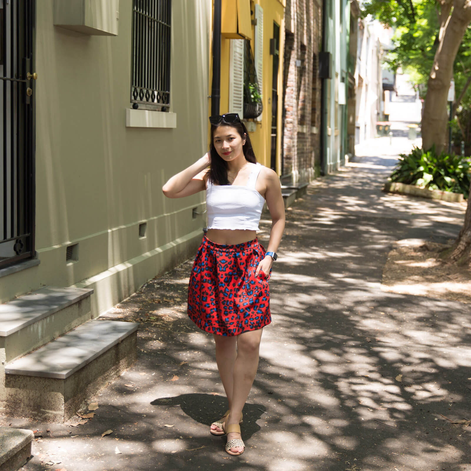 A woman standing on a footpath next to steps that lead up to the front door of houses. The area is shaded by trees. The woman has dark hair, and is wearing a white top with a red skirt with blue leopard sports. She has one hand in the pocket of her skirt and the other reaching up behind her head with her elbow out to the side