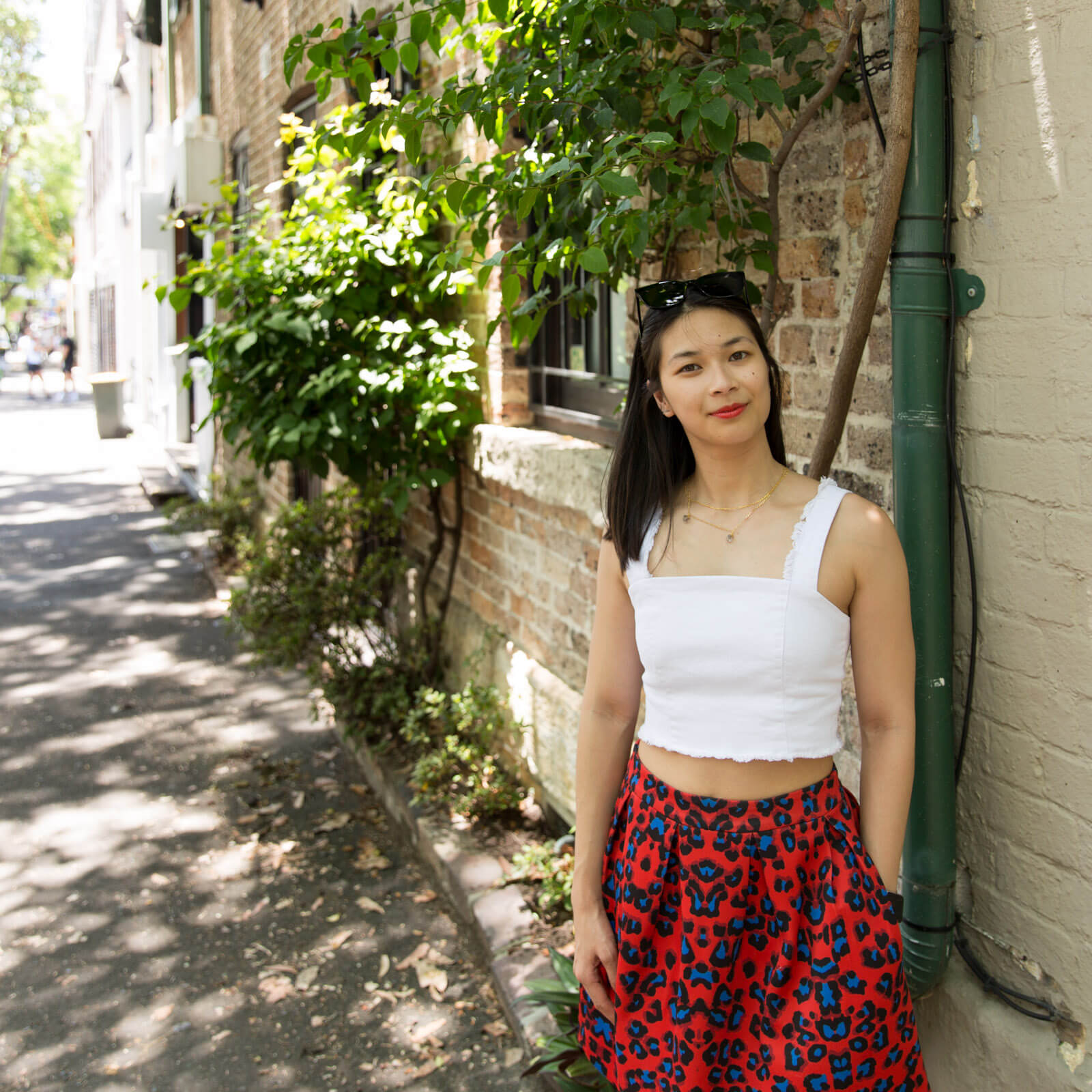 The same woman from other images on this page, in the same outfit. She is leaning against a green painted pipe attached to a brick wall. Just behind her is a window in a brick building and there are leaves and vines growing around the window.