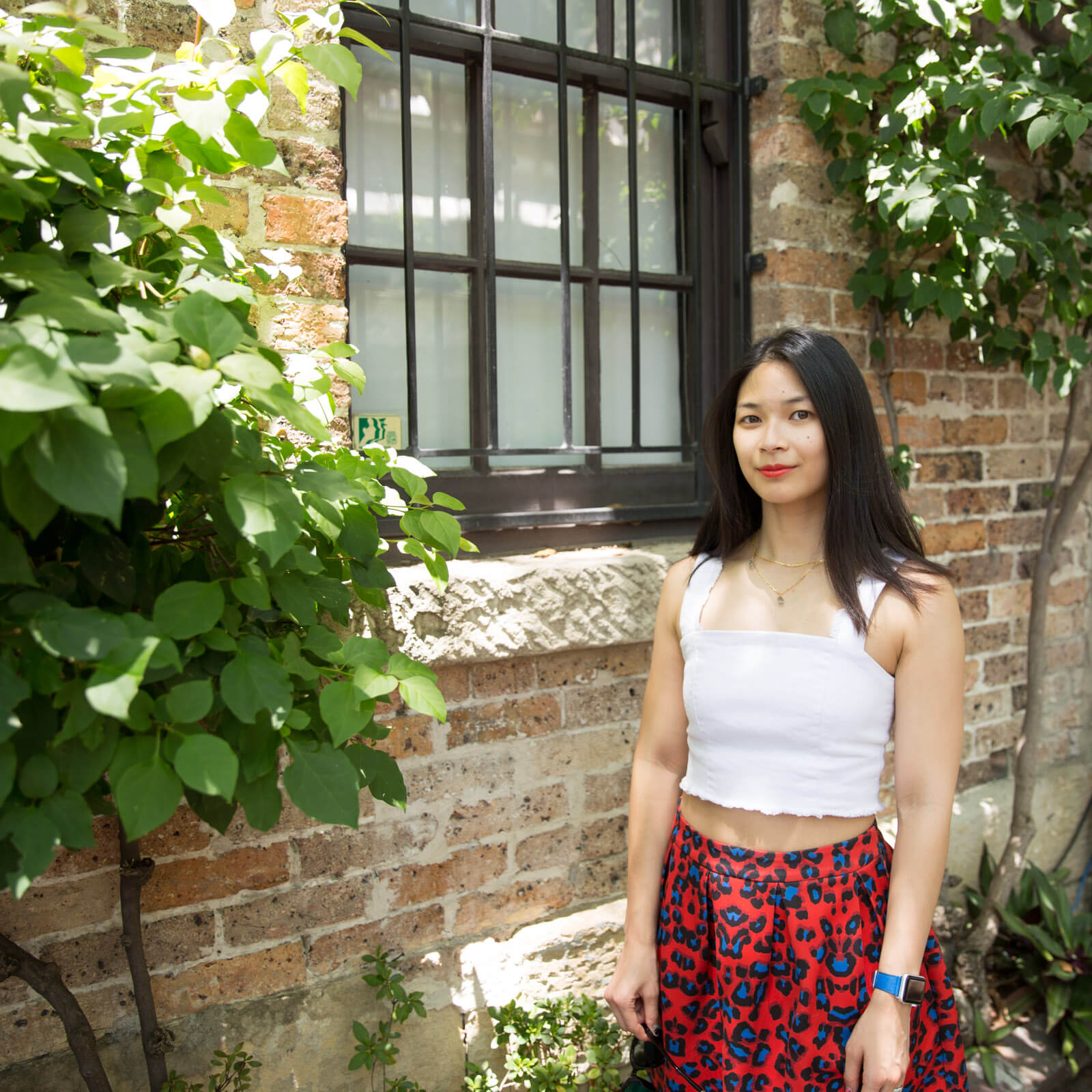 The same woman from other images on this page, in the same outfit. She is standing by a closed window in the brick wall of a building. There are some growing vines on the wall.