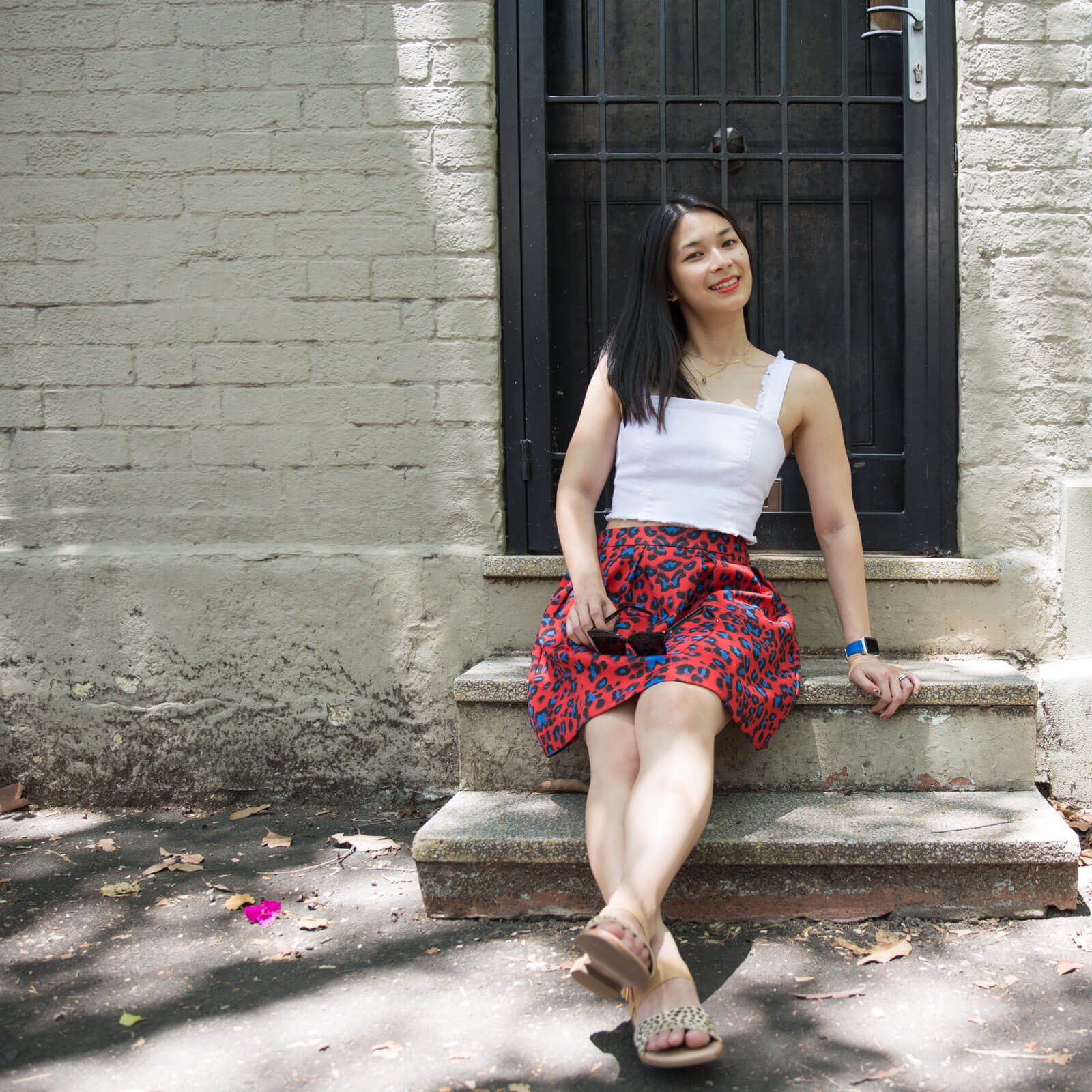 A woman with dark hair, wearing a white top with a red skirt with blue leopard sports, is sitting on the second step up from the ground, with a black door just behind her, in a grey brick building. The woman has her legs outstretched in front of her and towards the came and a hand resting to her side.