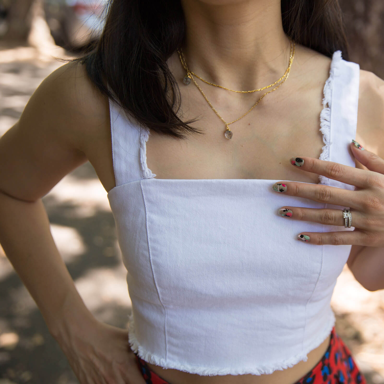 A closeup photo of a woman’s torso – the same woman featured in other photos on this page. She has a hand to her upper chest and the artwork on her nails is visible – subtle green with patches of green, black and pink. She has a double-layered chain necklace around her neck, from which two grey-coloured herkimer diamonds hang.