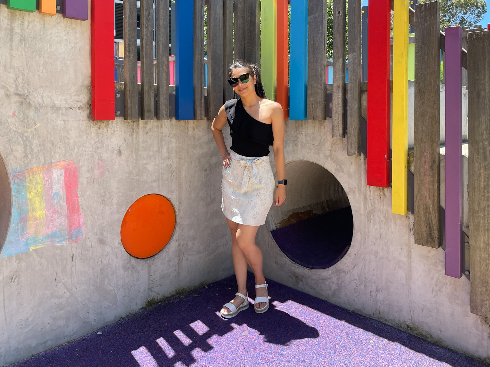 A woman wearing a black top with one shoulder, a floral white skirt and white platform sandals. She has black sunglasses on and has a hand on her hip. She is standing in front of a concrete children’s play structure with colourful wooden panels.